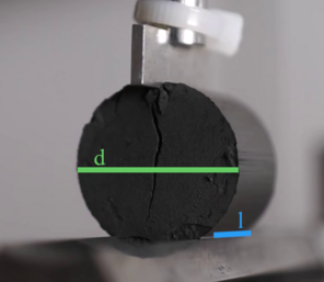 The image shows a compressed, granular graphite cylinder during break up. During break up a sudden decrease of the tensile strength can be seen. The maximum tensile stress is measured with a scale beneath the sample and provides the tensile strength of the material.